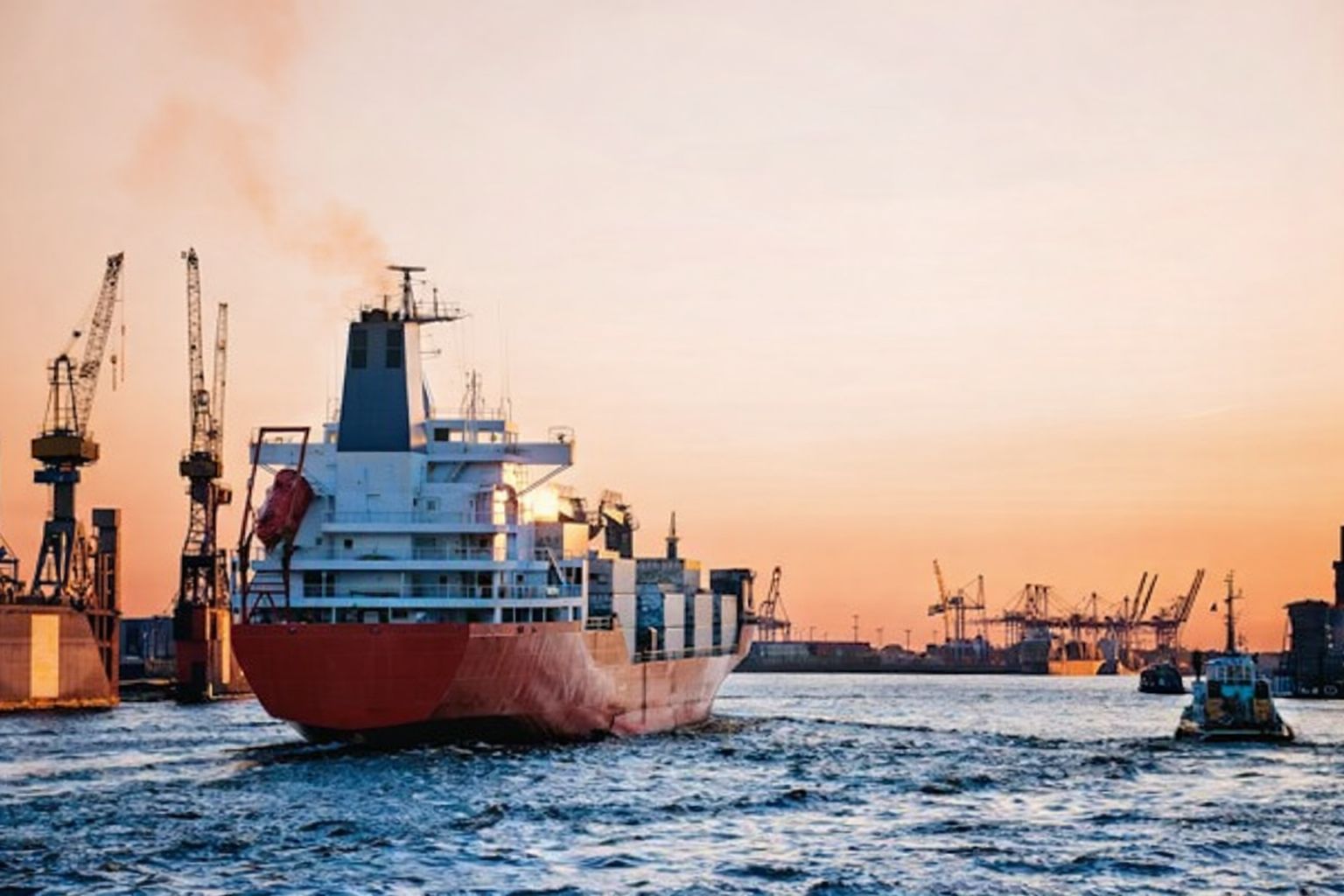 Lastfartyg på vatten med hamnkranar och solnedgång i bakgrunden.